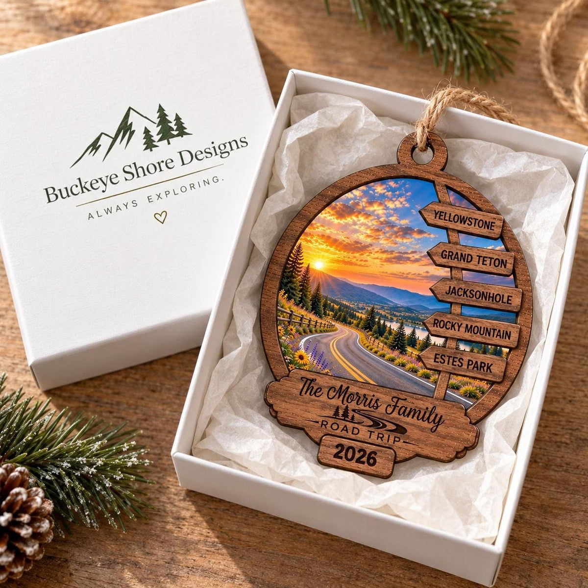 The wooden ornament lies inside an open white gift box featuring the Buckeye Shore Designs logo on the lid, resting on a wooden surface with a pine sprig.