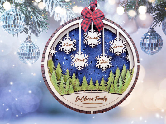 May include: A round wooden Christmas ornament features a winter forest scene with a dark blue night sky, green trees, and snowflakes with family names: