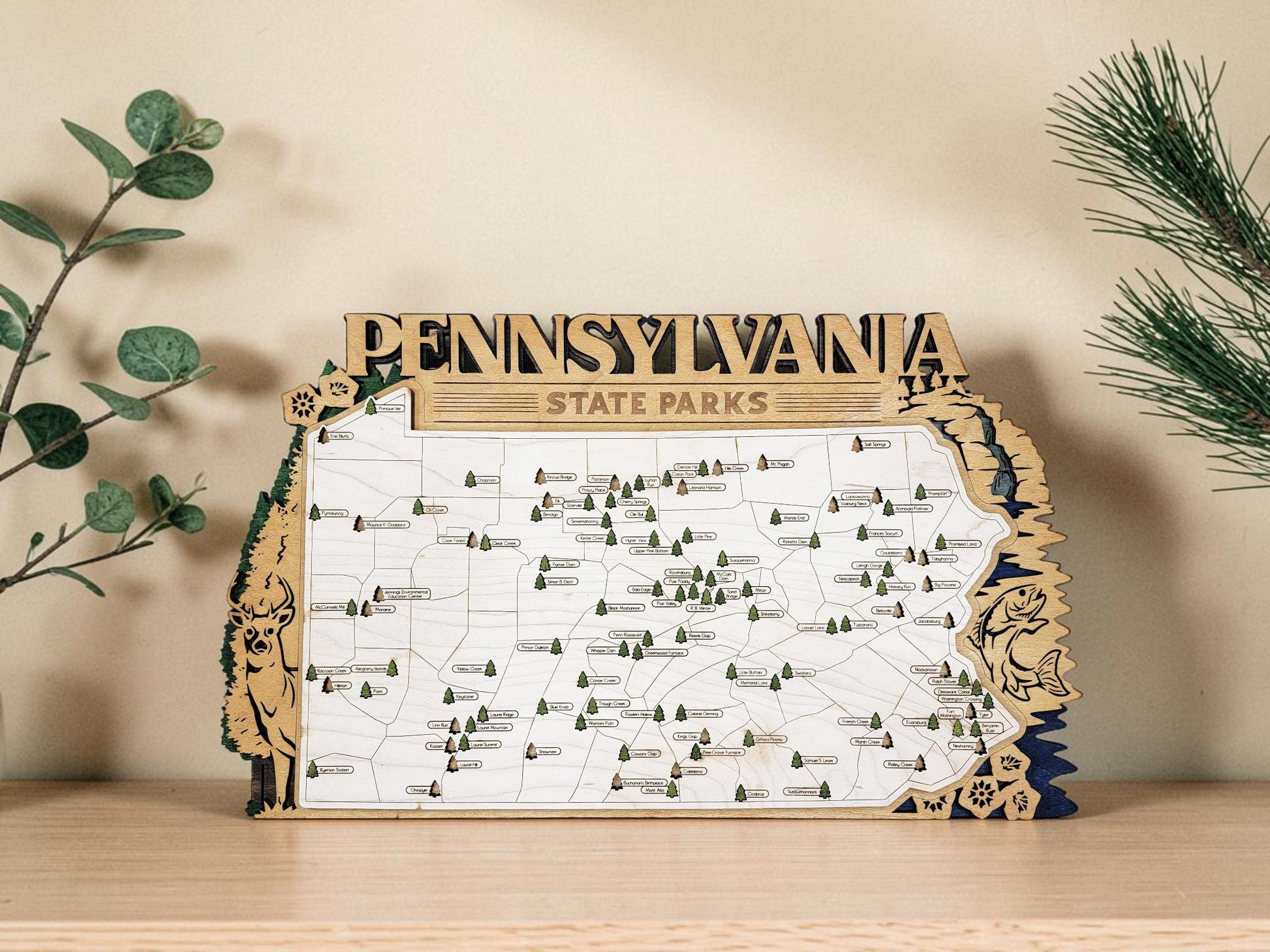 May include: Wooden map of Pennsylvania State Parks. The map is in the shape of the state, with the name "PENNSYLVANIA" at the top. The map has small green tree icons and is surrounded by a decorative border with a deer and fish.