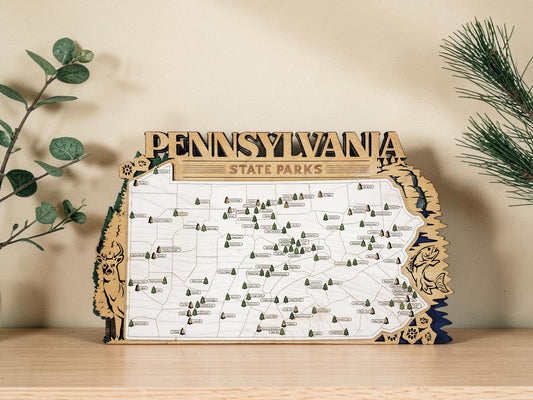 May include: Wooden map of Pennsylvania State Parks. The map is in the shape of the state, with the name "PENNSYLVANIA" at the top. The map has small green tree icons and is surrounded by a decorative border with a deer and fish.