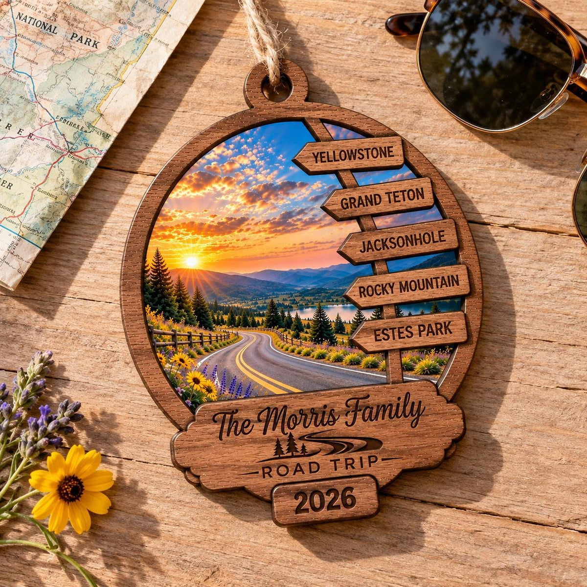 The personalized wooden ornament rests on a wooden table next to a map, sunglasses, and yellow wildflowers.